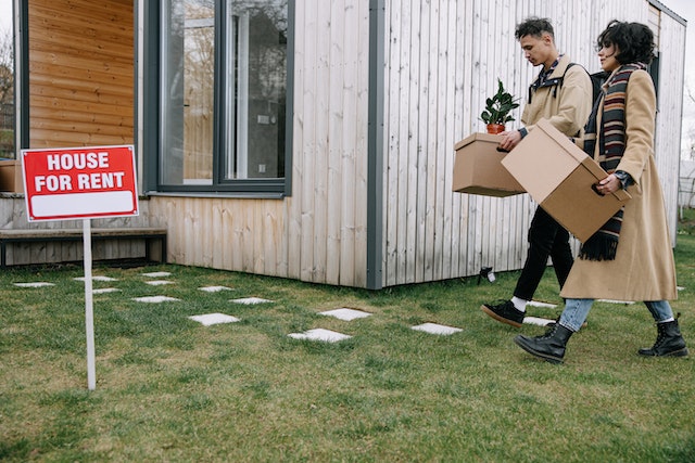 Two people carrying moving boxes into a house with a For Rent sign outside of it